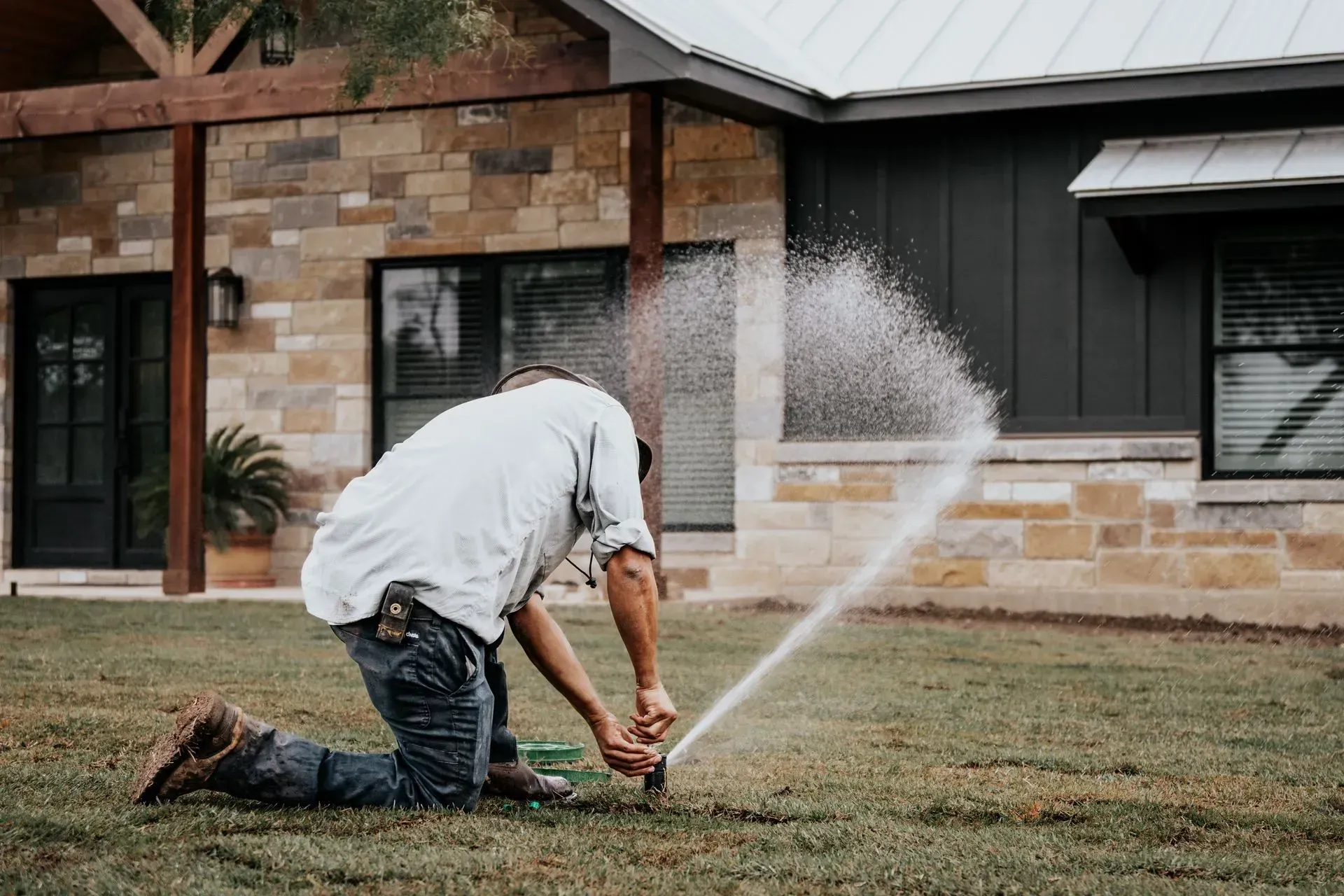 Man adjusting a sprinkler in front of a house, water spraying.