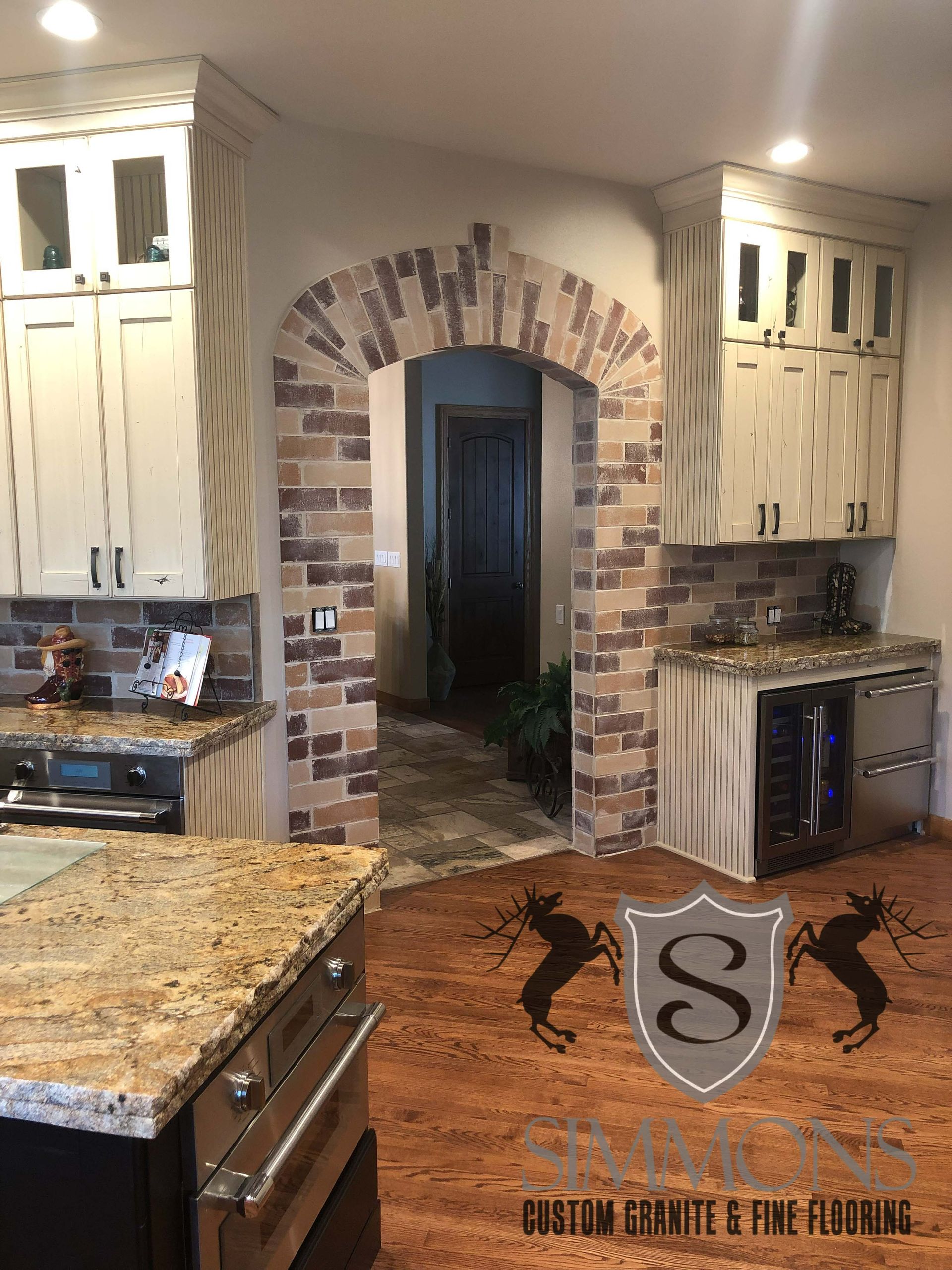 Kitchen with granite island, white cabinets, brick archway, and dark wood floor.
