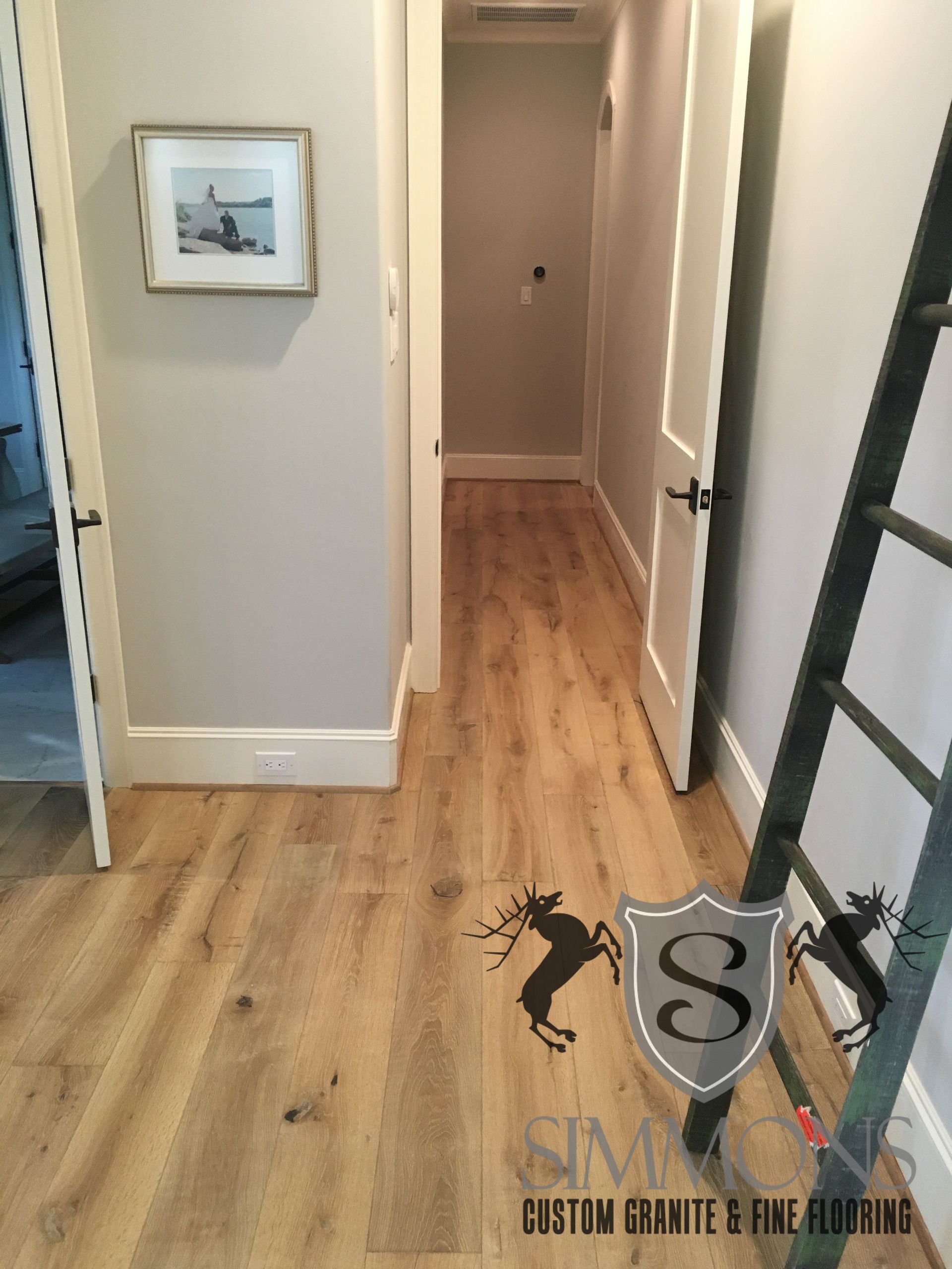 Hallway with light wood floors, beige walls, framed art, and a dark staircase railing.