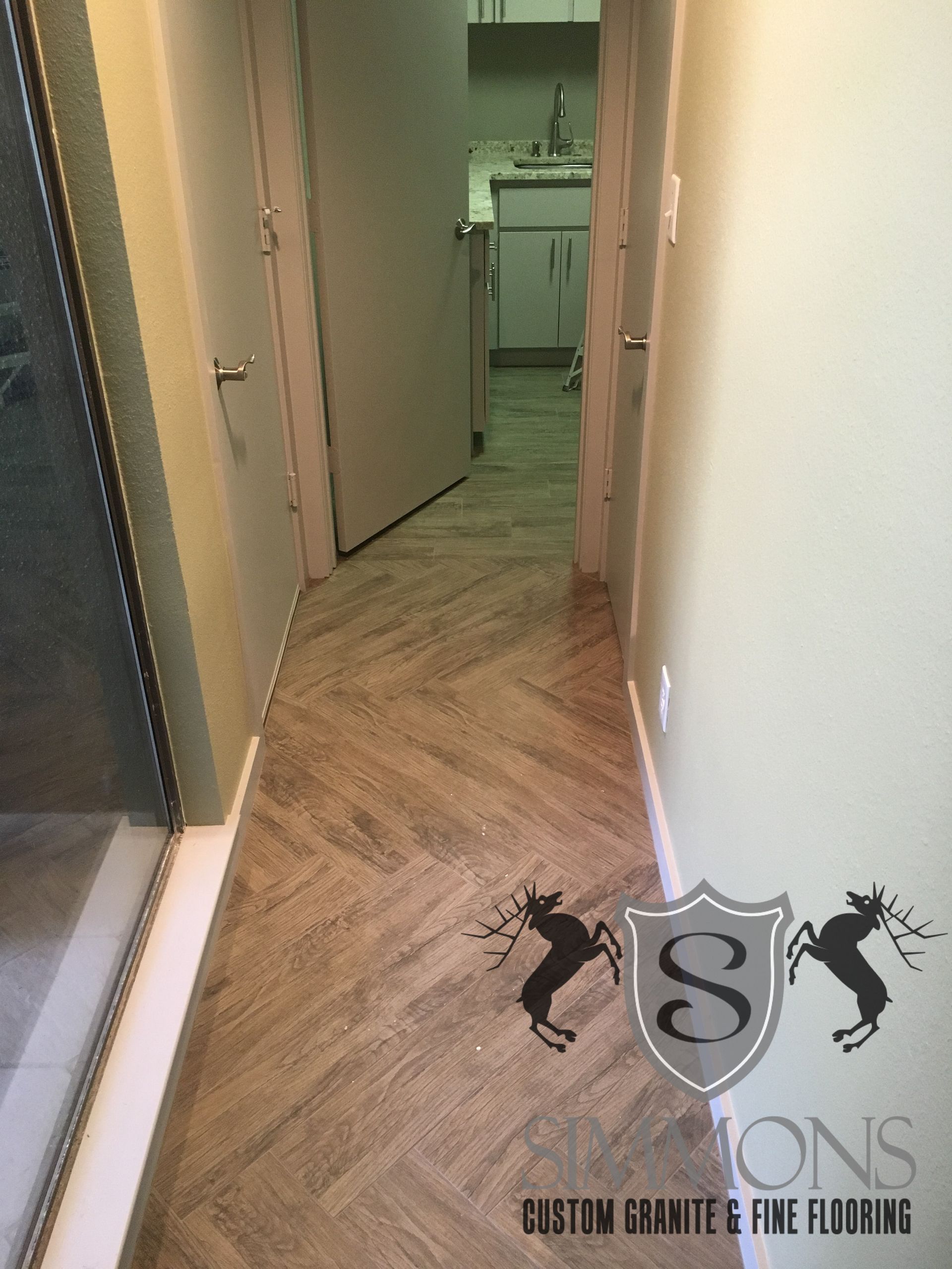 Narrow hallway with herringbone wood floor, open doorways, and a kitchen visible at the end.