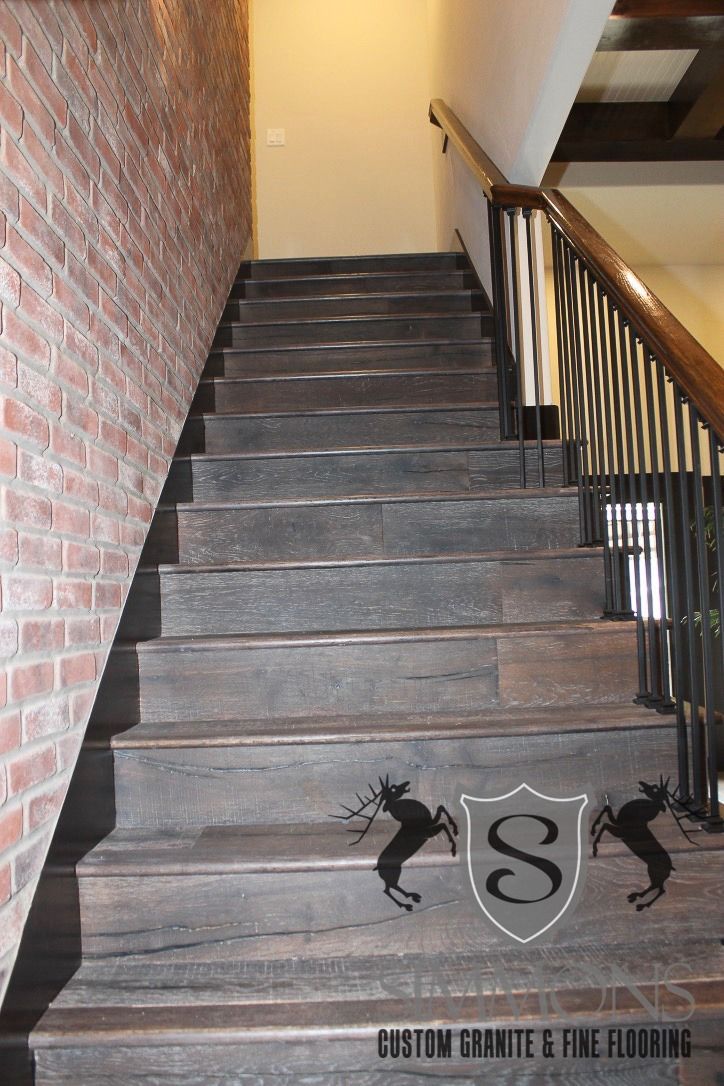 Narrow indoor staircase with gray steps, brick wall on left, and black railing on right.