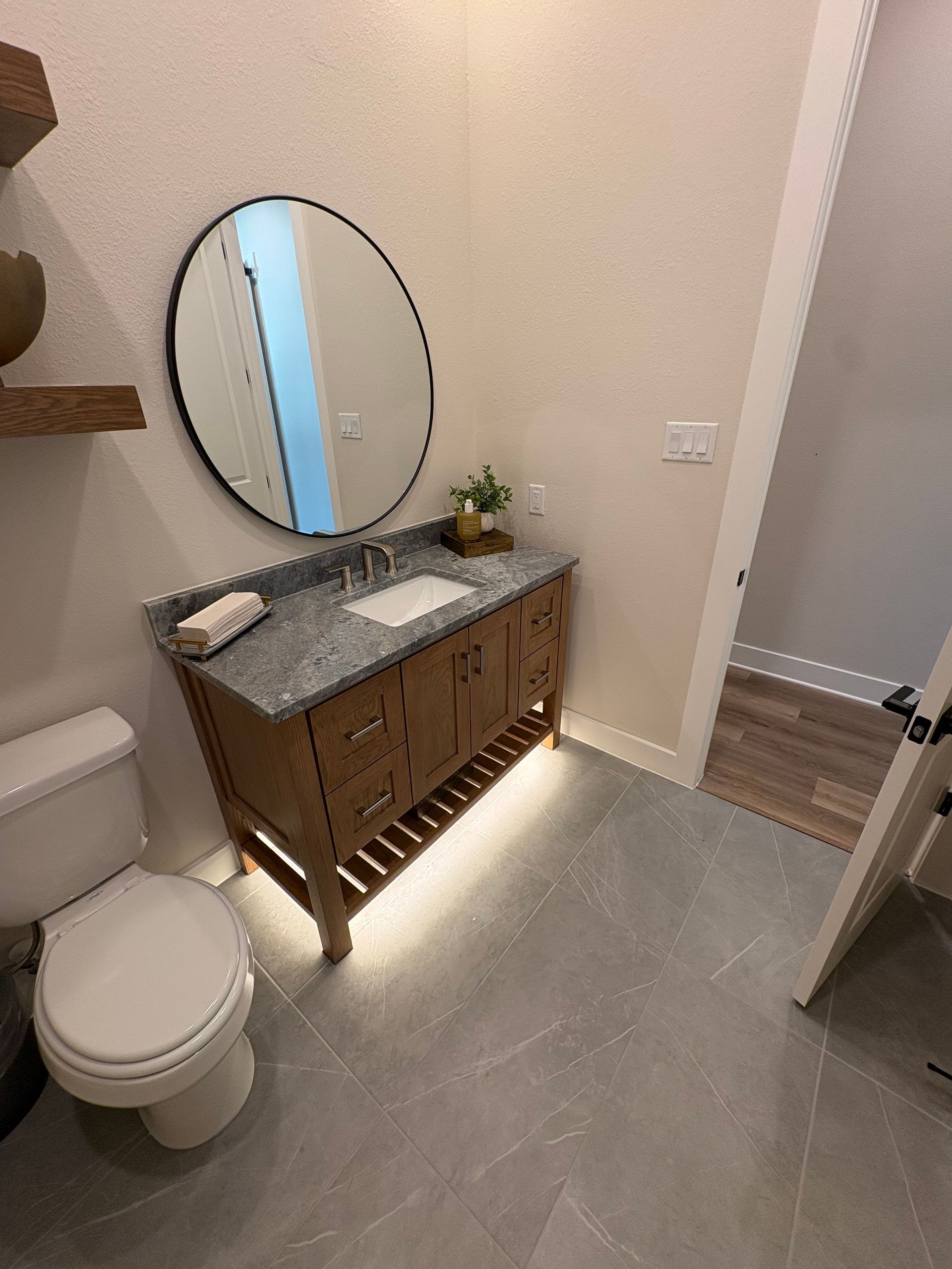 Modern bathroom with floating wood vanity, round mirror, toilet, and tiled floor; warm under-cabinet lighting