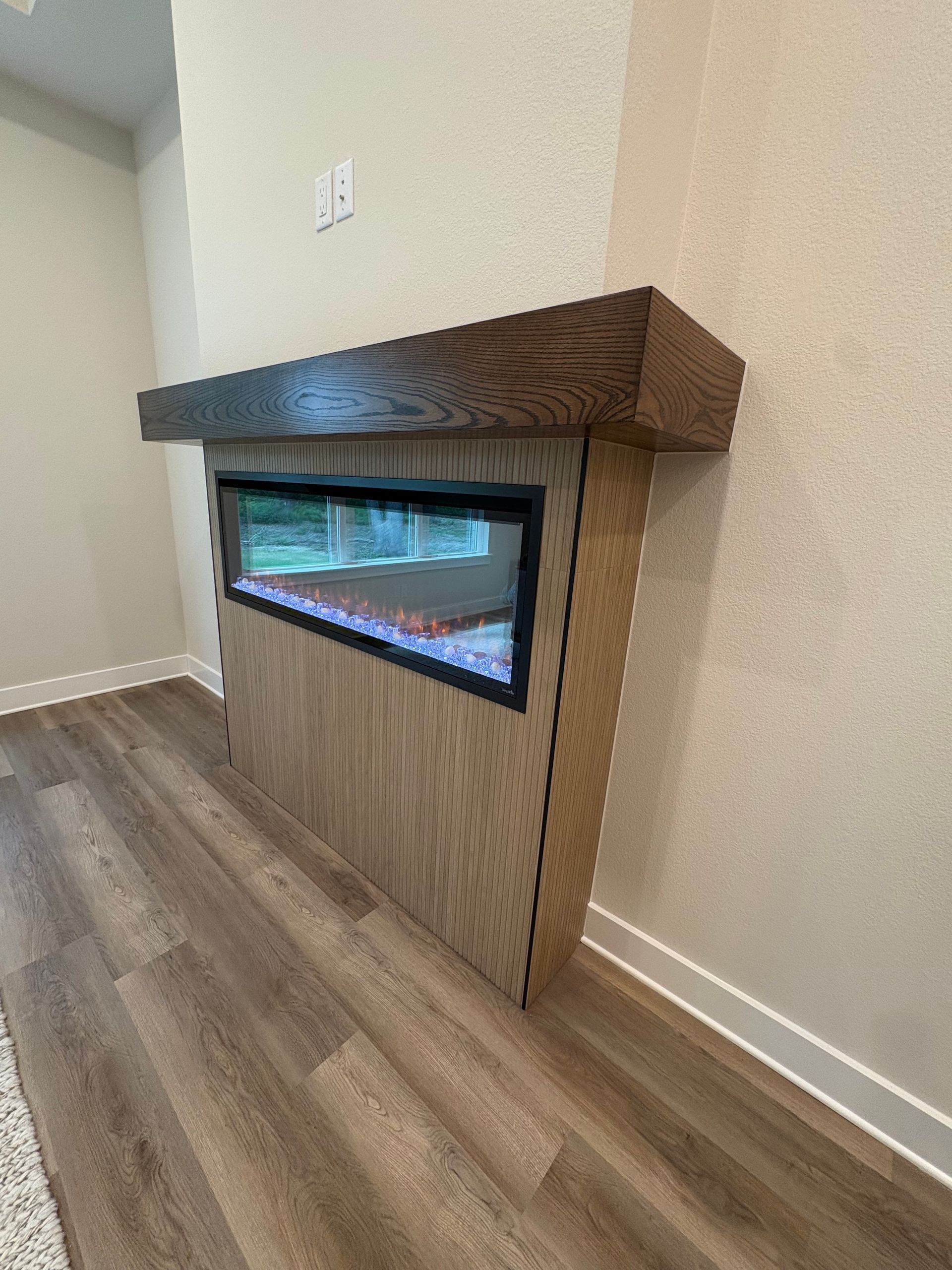 Modern electric fireplace with dark wood mantel in a light beige room, next to wood-look flooring.