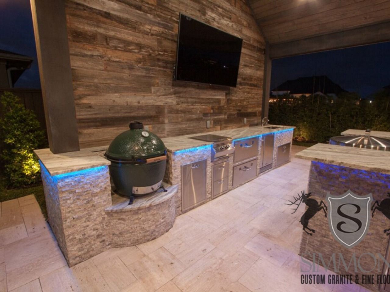 Outdoor stone patio with blue-lit built-in counters, green grill, wall-mounted TV, and a hot tub at sunset