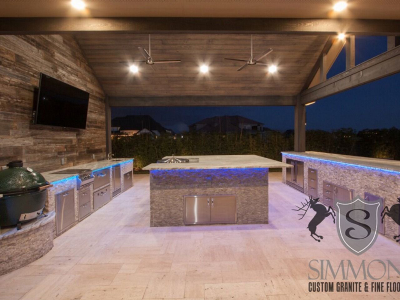 Covered outdoor kitchen with stone counters, TV, bar stools, and a lit pool at dusk
