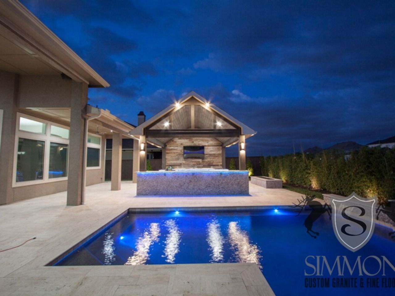 Modern backyard patio with lit pool, stone cabana, and blue dusk sky