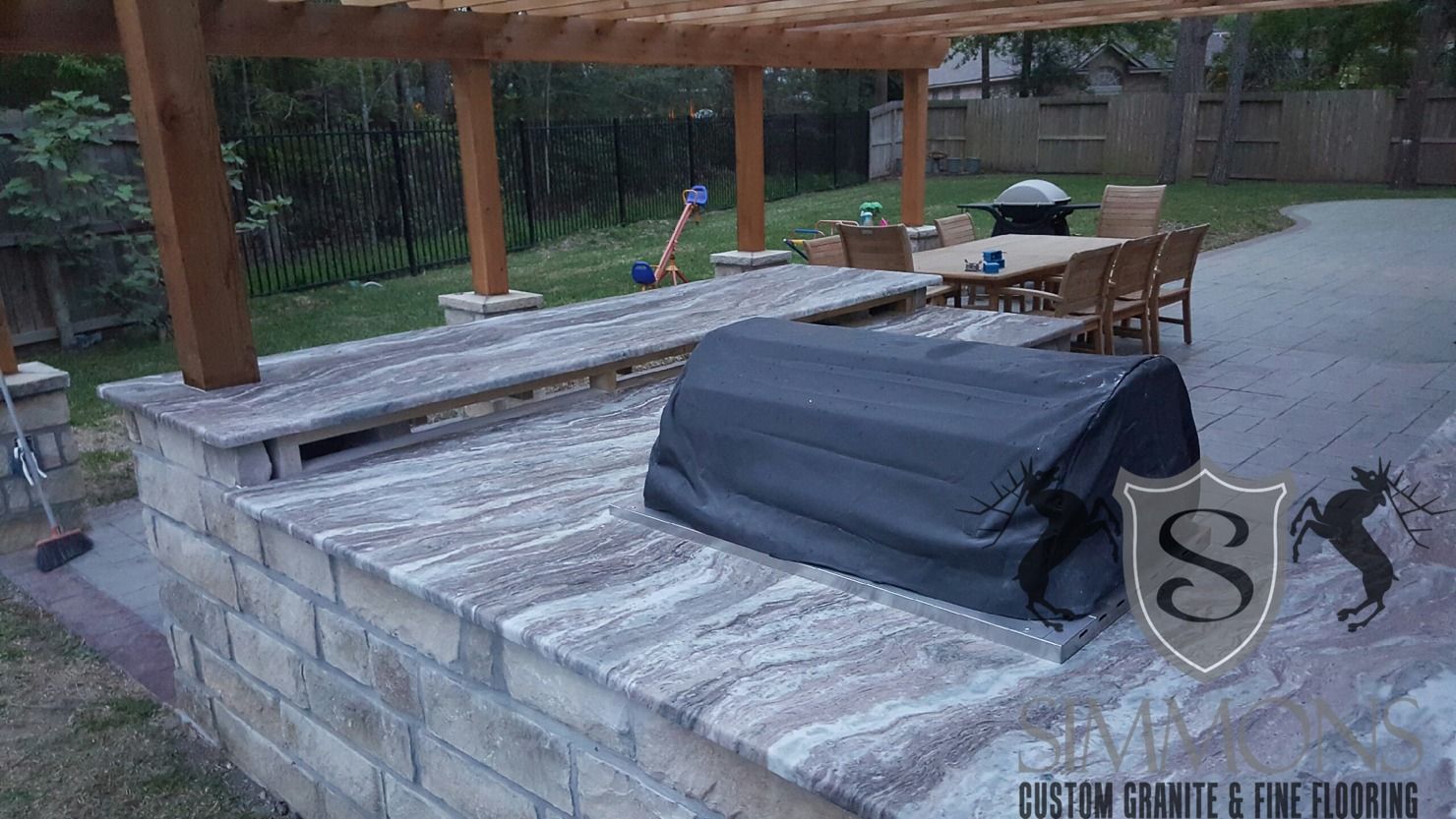 Stone outdoor patio with a covered grill, dining table, and wooden pergola by greenery
