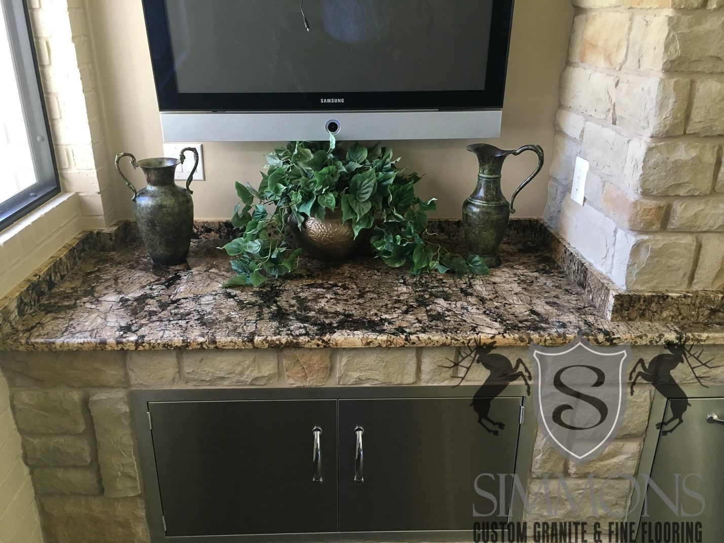 Stone bathroom vanity with granite top, silver sink, decorative plants, and wall-mounted TV above.