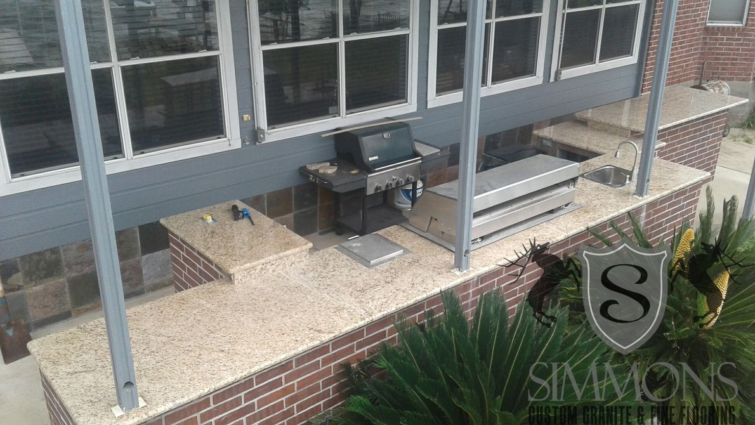 Brick patio with outdoor grill and table beside a gray house, viewed from above.