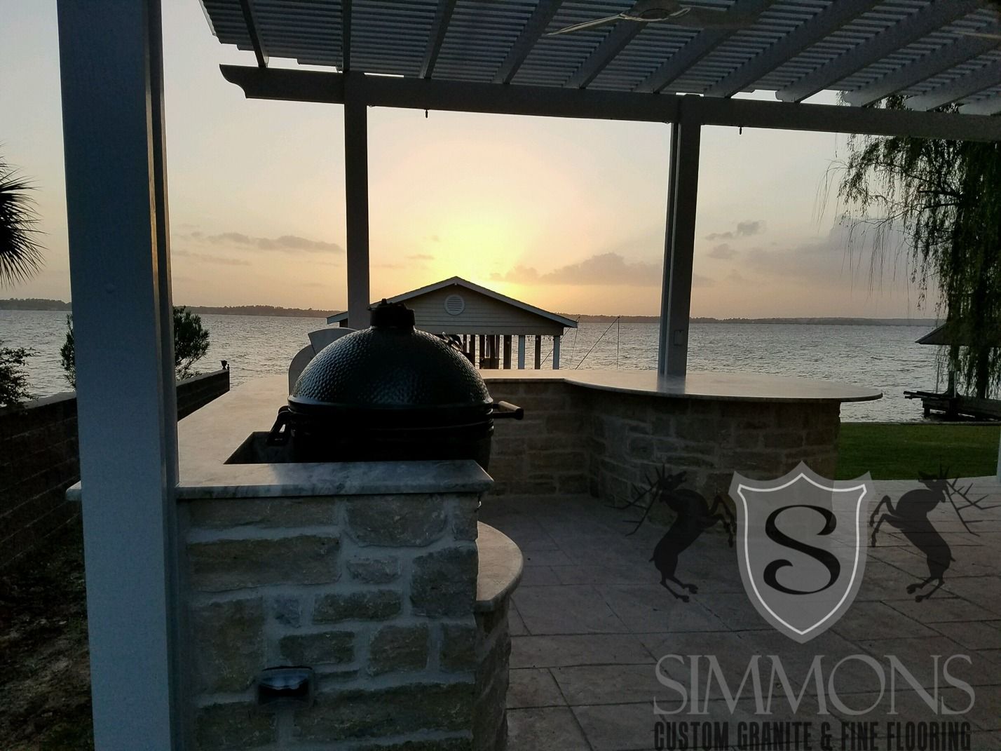 Sunset over a waterfront patio with stone pillars, a grill, and a small dock over calm water