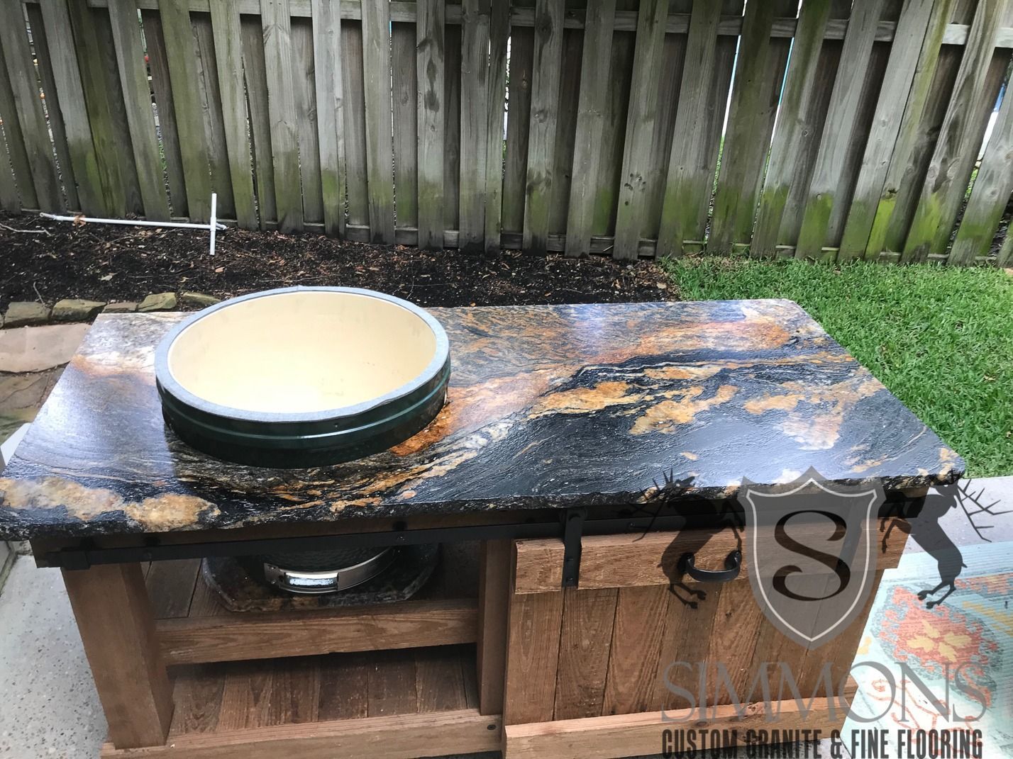 Outdoor wooden table with a black-and-gold bowl on top, in a fenced backyard.
