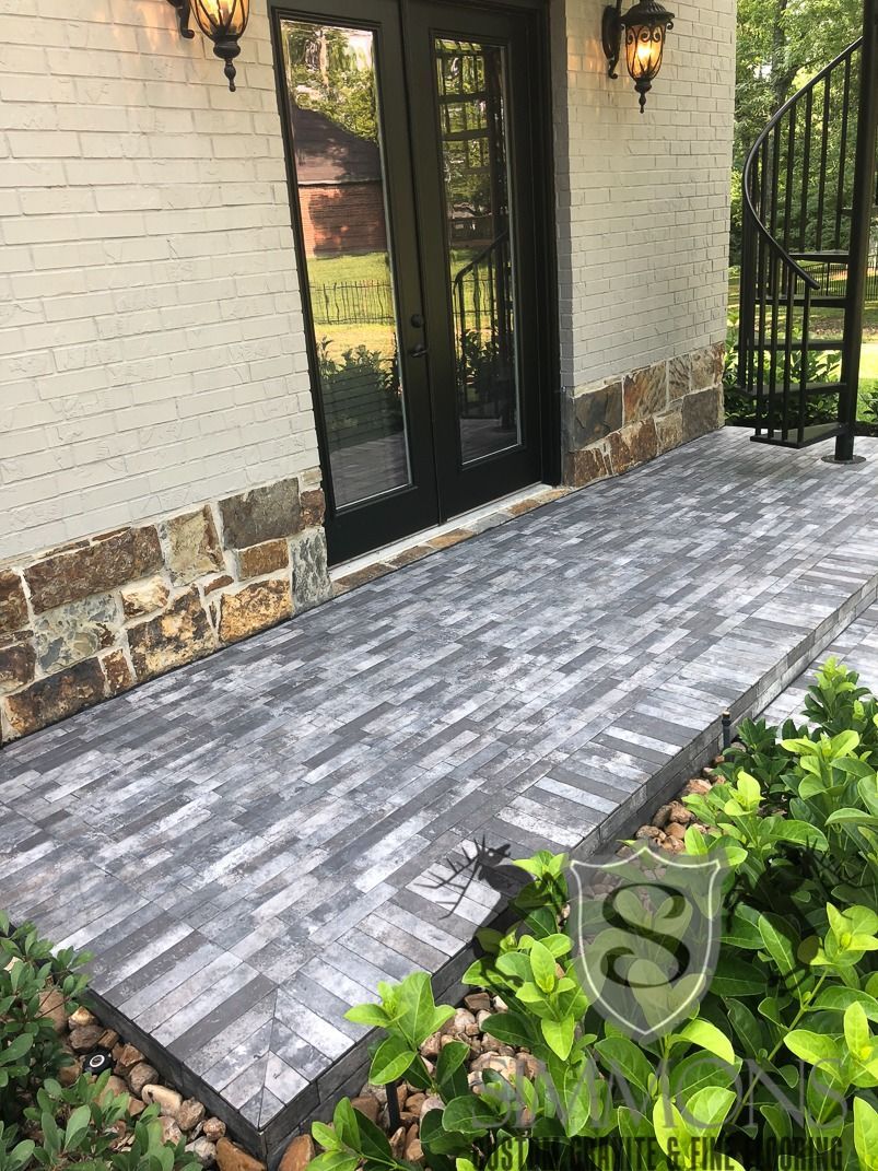 Brick front porch with gray paver tiles, black double doors, and wall lanterns beside a landscaped garden.