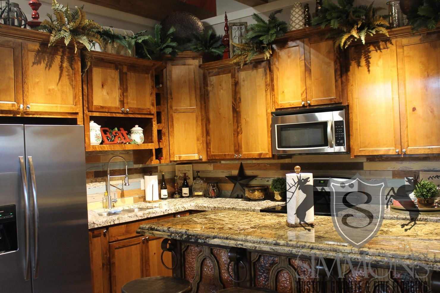 Warm kitchen with wooden cabinets, granite countertops, stainless fridge, and microwave under decorative greenery