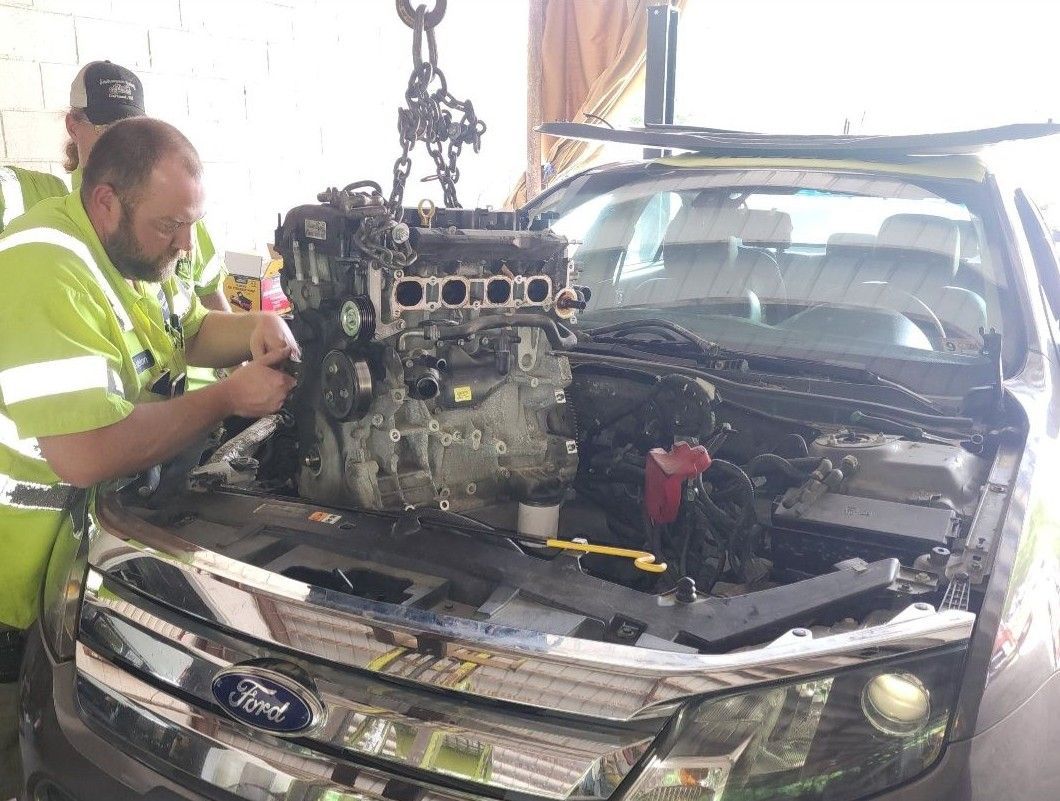 Two people in neon vests lifting a car engine out of a Ford hood. A crane holds the engine in place.