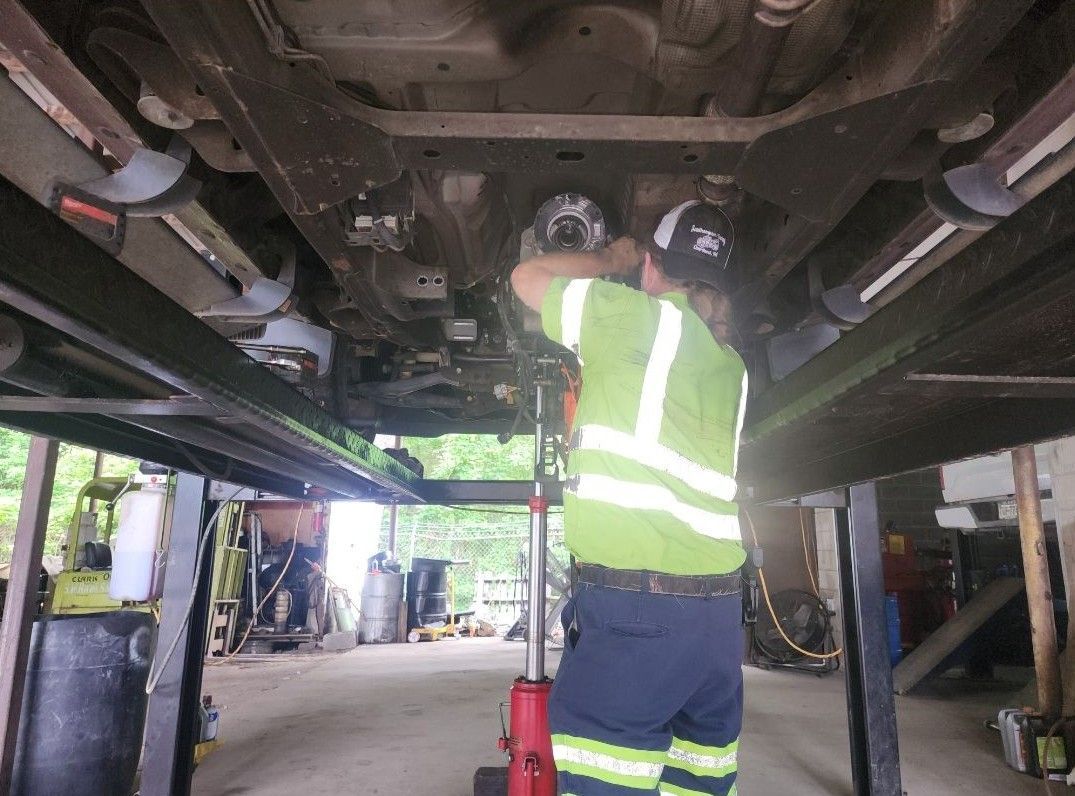 Mechanic working under a car on a lift; wearing high-vis vest and holding a tool. Shop setting.