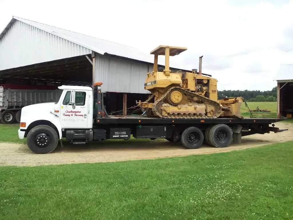 White flatbed truck carrying a yellow bulldozer on a grassy field, near a barn.