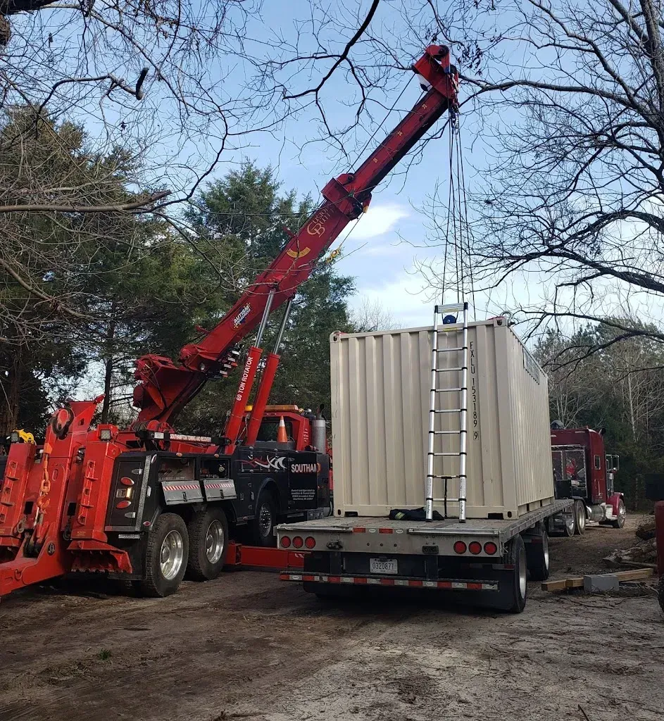A red tow truck with a crane lifting a rectangular container onto a trailer in an outdoor setting.