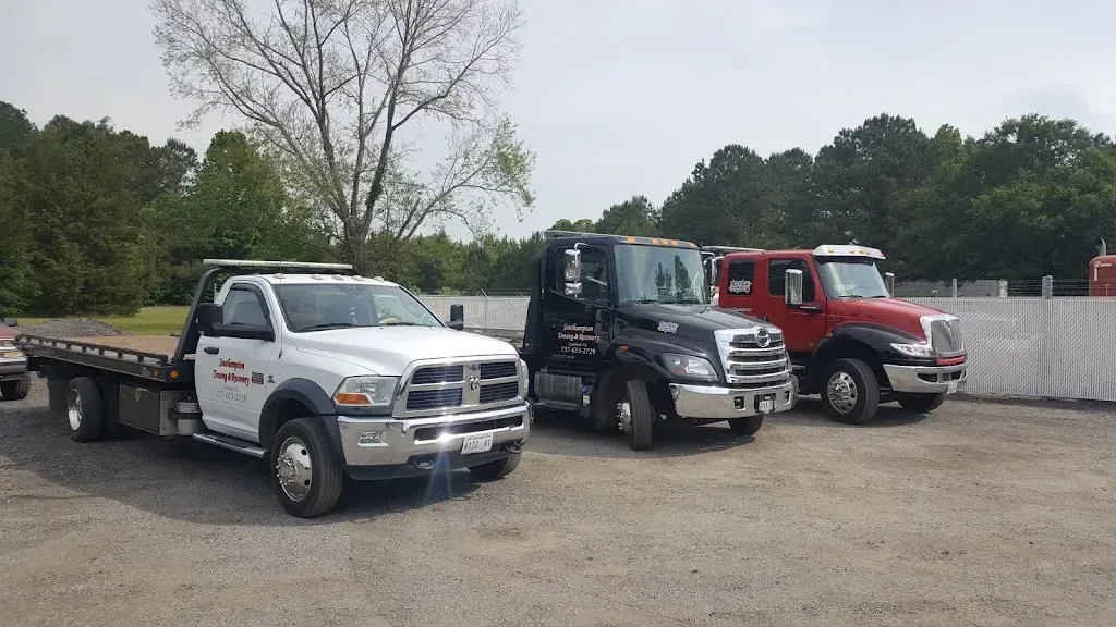 Three tow trucks parked on gravel, white, black, and red, in front of trees under a cloudy sky.