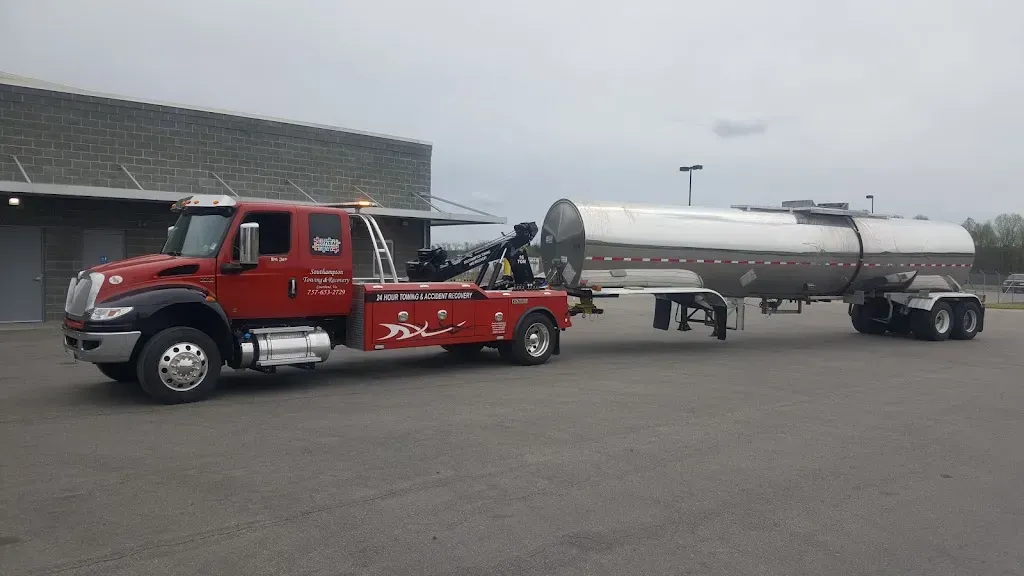 Red tow truck towing a large silver tanker trailer in a parking lot on a cloudy day.