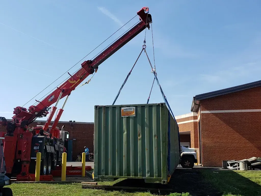 A red crane lifting a green shipping container; blue sky, brick building.