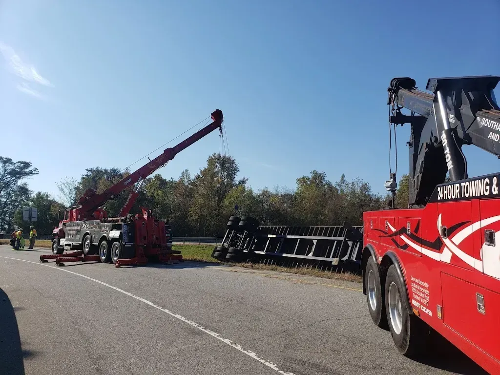 Two red tow trucks at a roadside accident. A semi-trailer is on its side. Trees and a clear sky in the background.