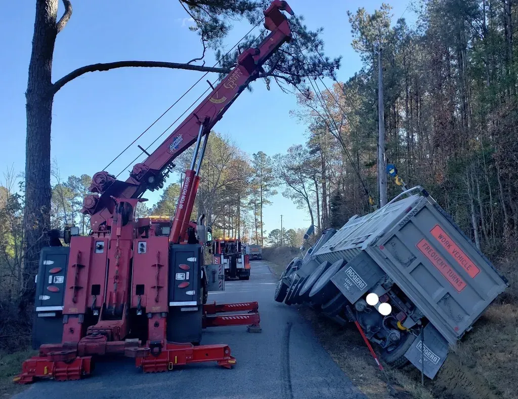 A red tow truck attempting to upright a tilted gray freight car on a road, trees and blue sky background.