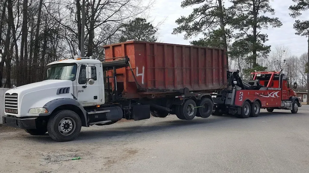 White dump truck with a red dumpster being towed by a red tow truck on a road.