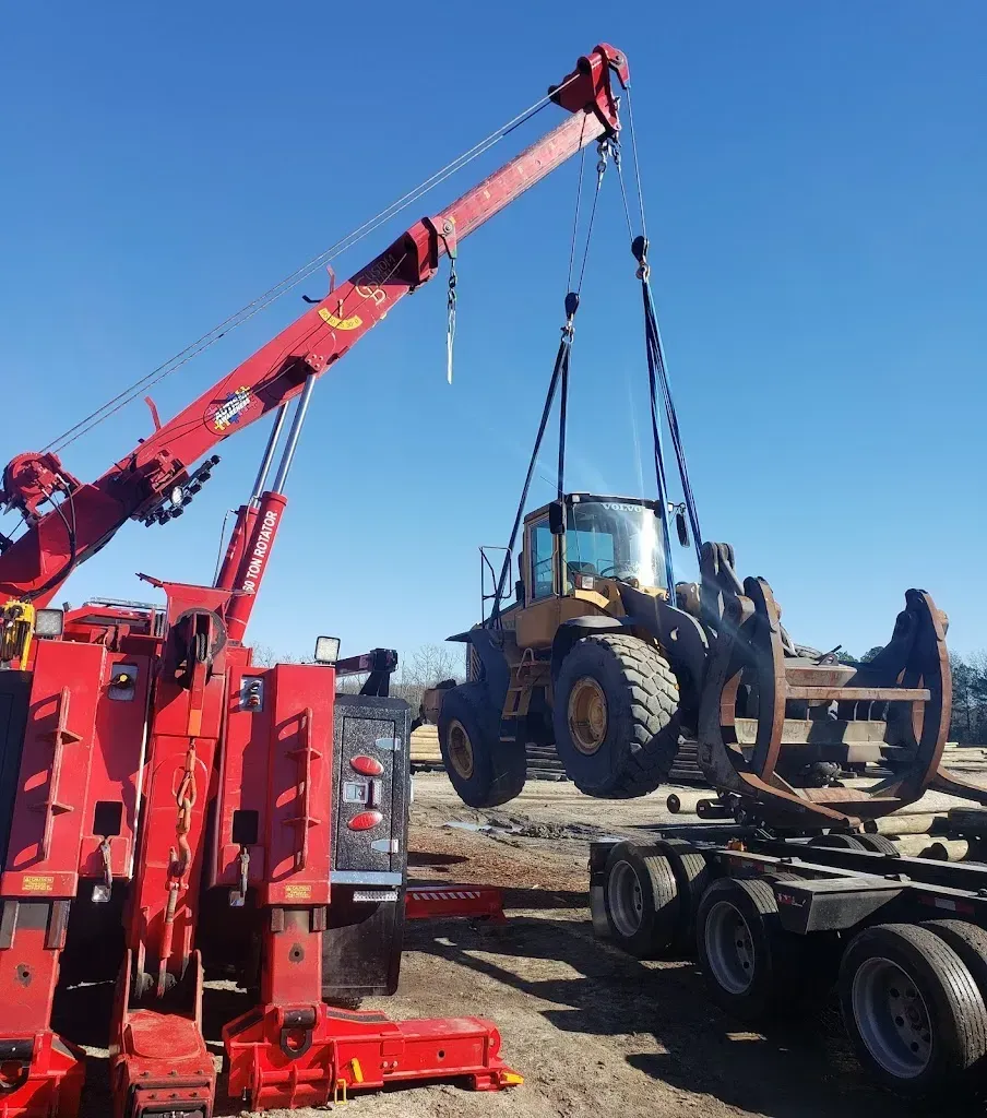 Red tow truck lifting a yellow front-end loader with a grapple attachment onto a trailer on a sunny day.
