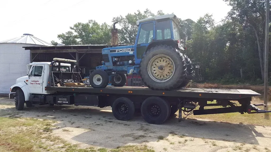 A blue tractor on a flatbed truck in front of a barn and silos on a sunny day.