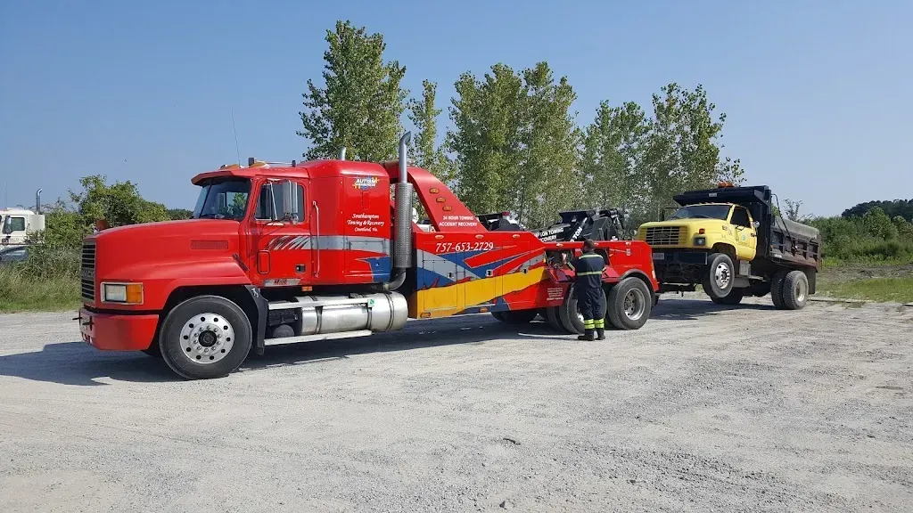 Red tow truck towing a yellow dump truck on a gravel road, blue sky background.