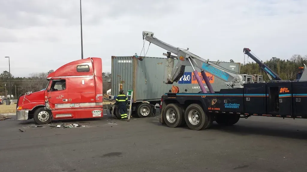 Red semi-truck damaged in collision being towed; a tow truck lifts the container.