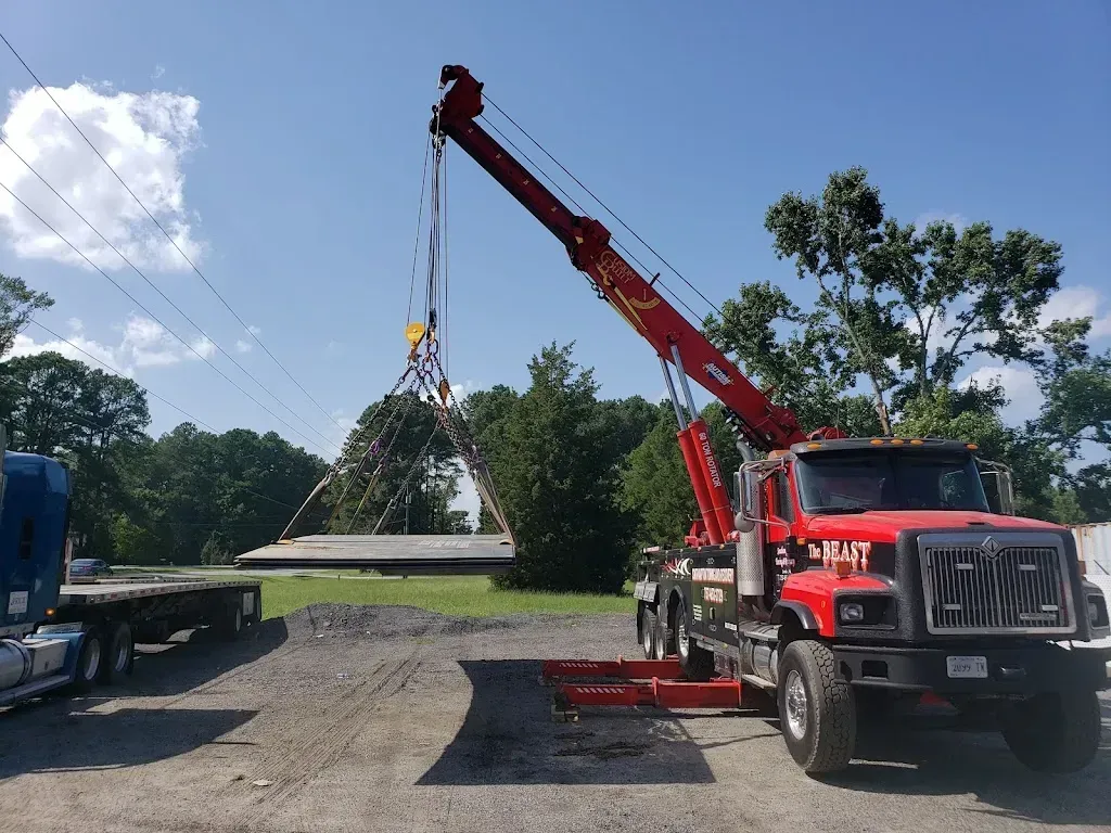 Red crane lifting concrete slabs, parked on gravel, with trees and blue sky in background.