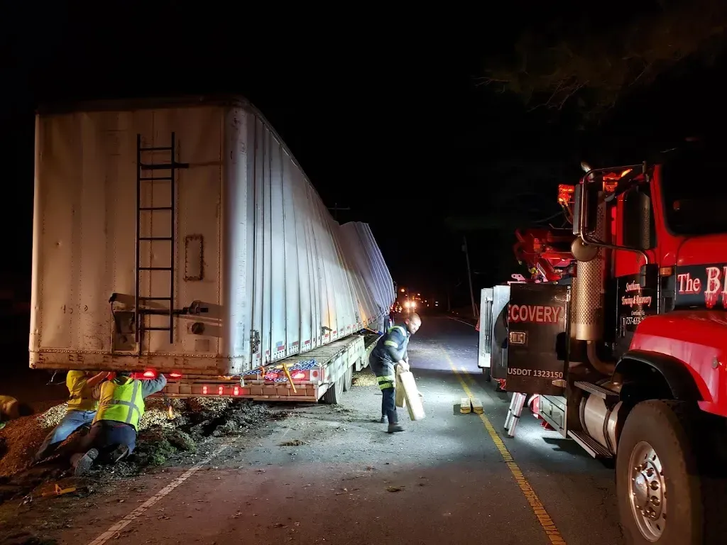 Semi-truck trailer overturned on road at night; tow truck, workers, emergency lights.