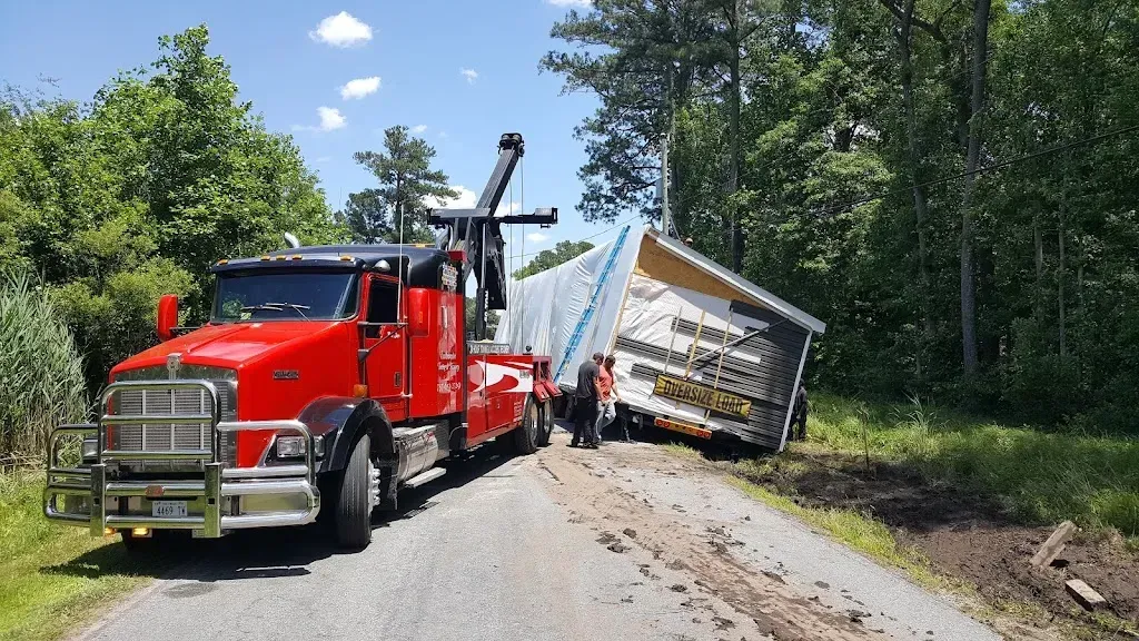 Red tow truck towing a tilted, partially damaged mobile home off a rural road; trees in the background.