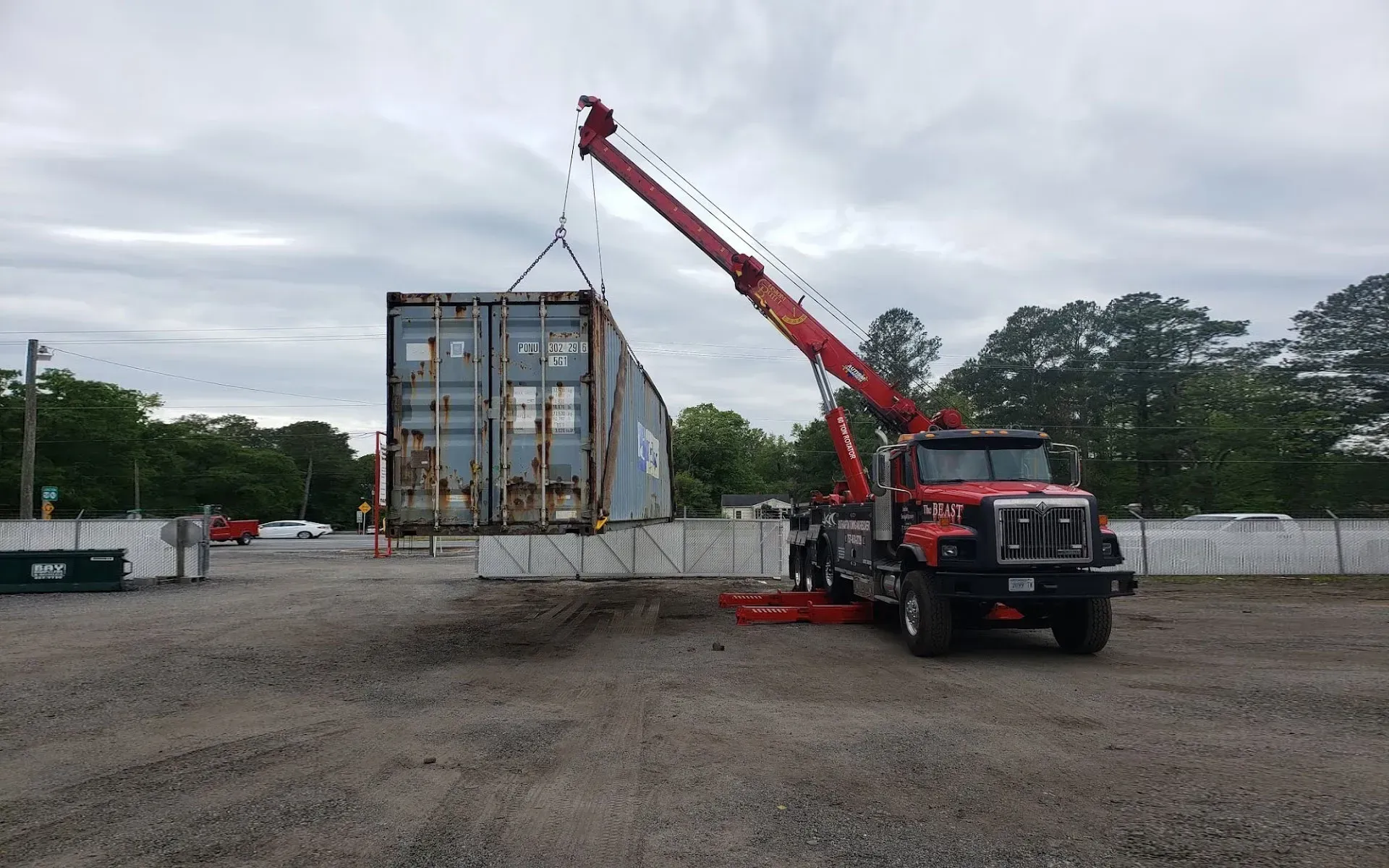 Red crane truck lifting a shipping container in an industrial area under a cloudy sky.