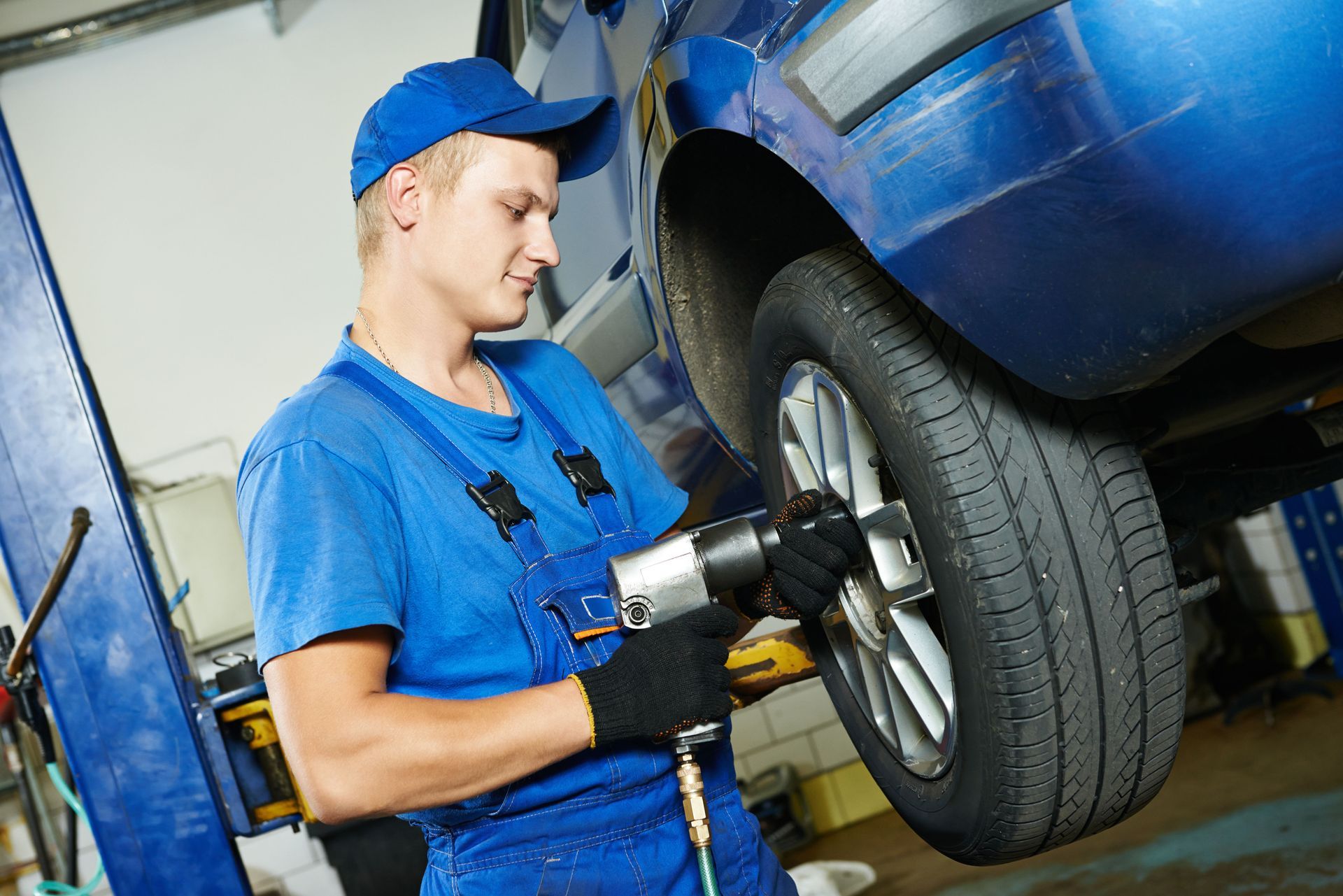Mechanic in blue uniform uses a pneumatic wrench on a car tire in a garage.