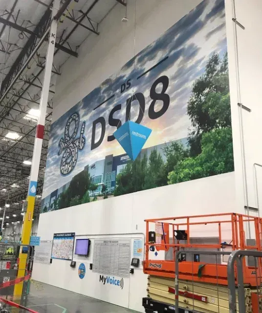 Large indoor industrial banner with “DSD8” and a blue-and-yellow logo above a warehouse wall and lift equipment.
