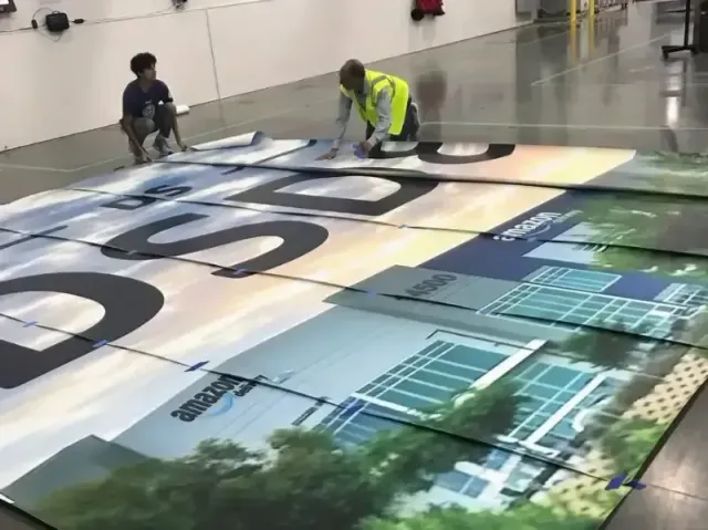 Two workers install a large colorful floor graphic in a warehouse-like space.