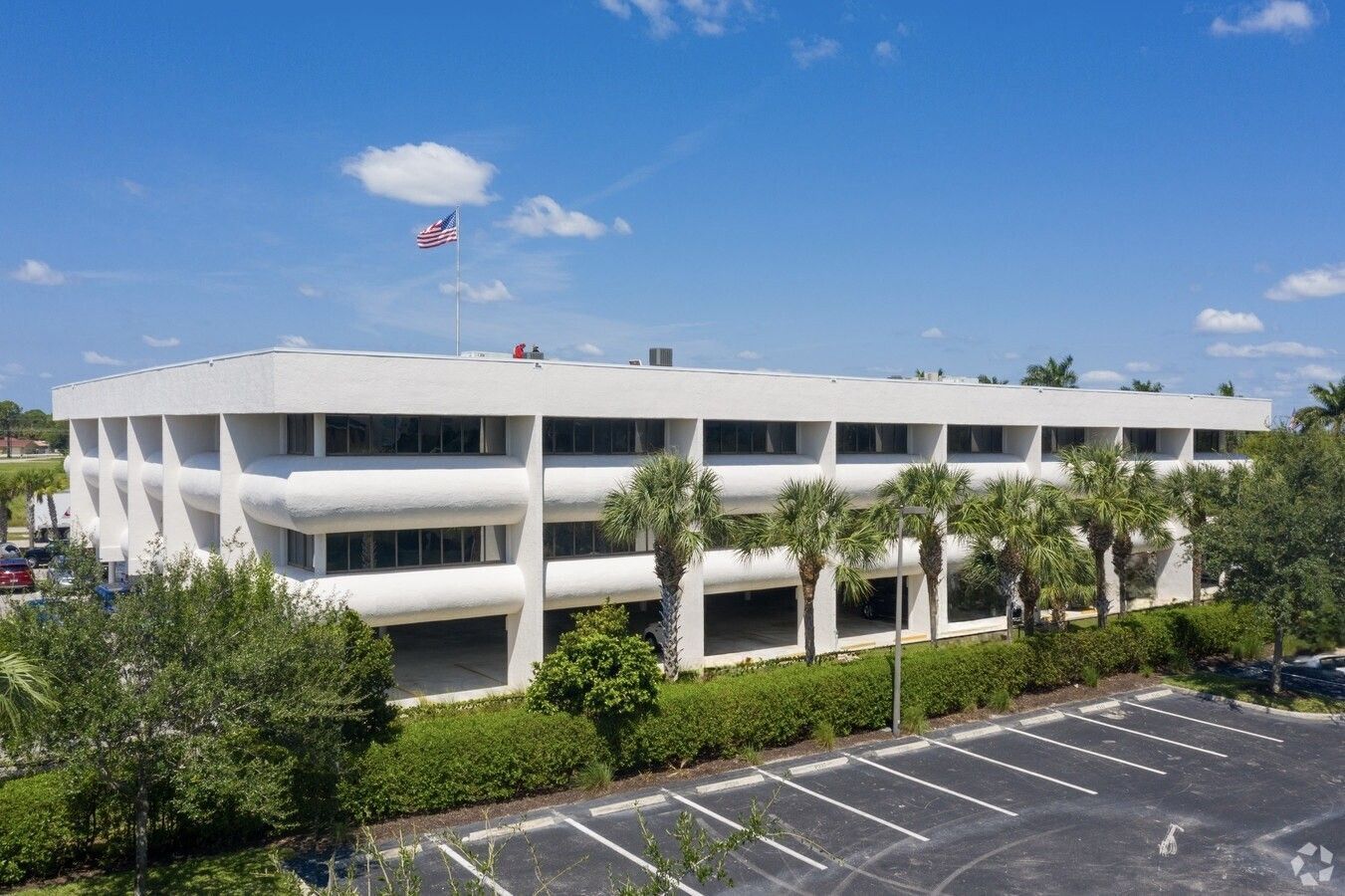 White office building with a U.S. flag on the roof, palm trees, and a parking lot.