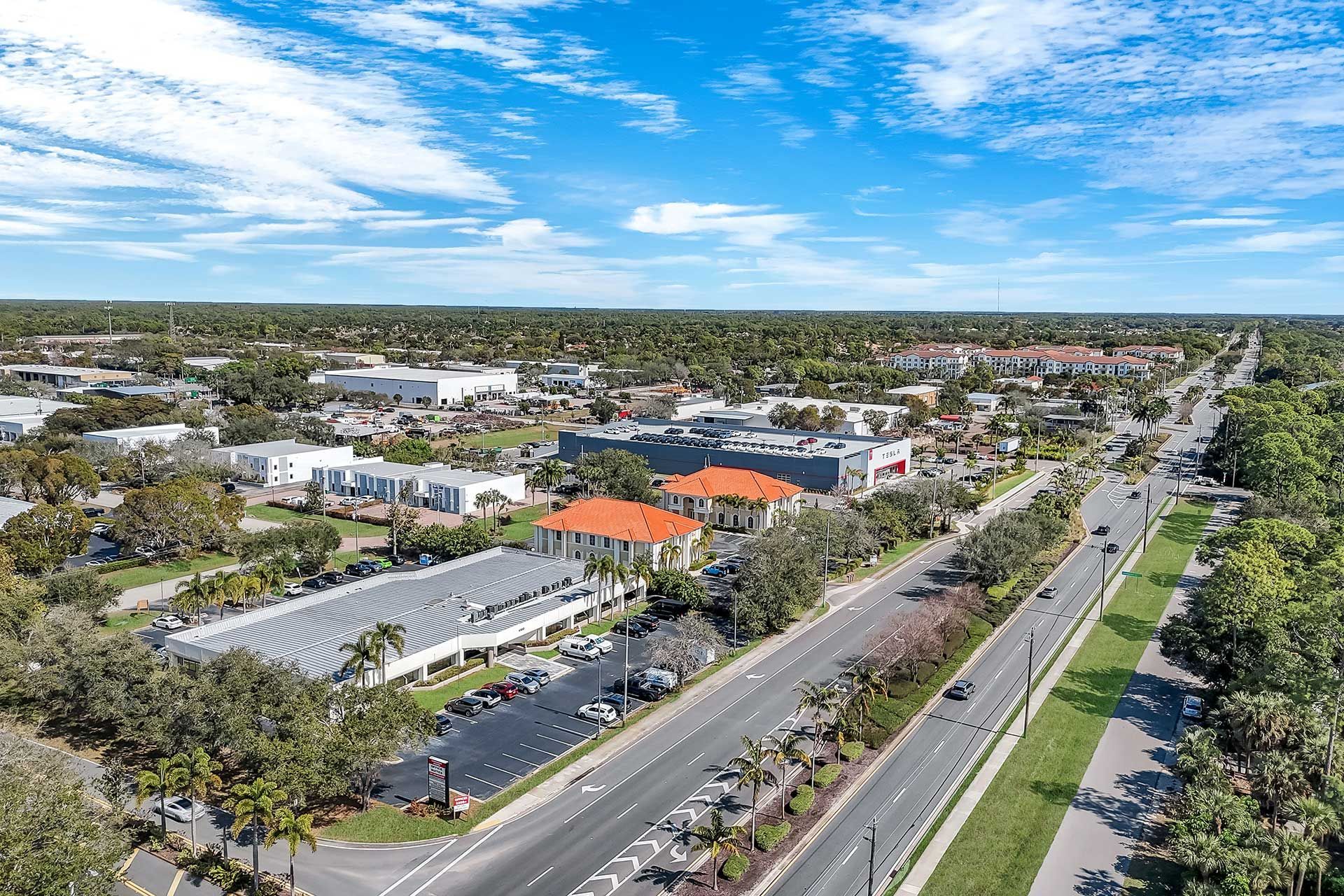 Aerial view of a suburban street lined with trees, buildings, and parked cars under a partly cloudy sky.