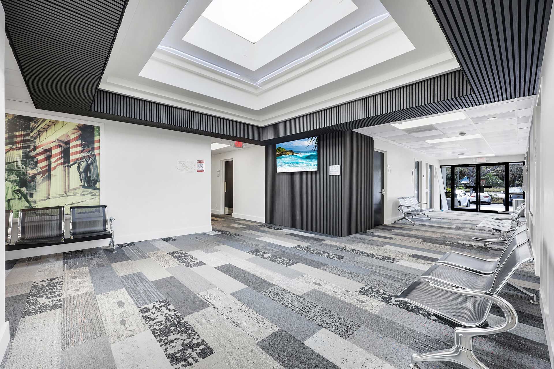 Reception area with gray, white, and blue patterned floor and ceiling panels. A screen shows a tropical beach. Chairs are present.