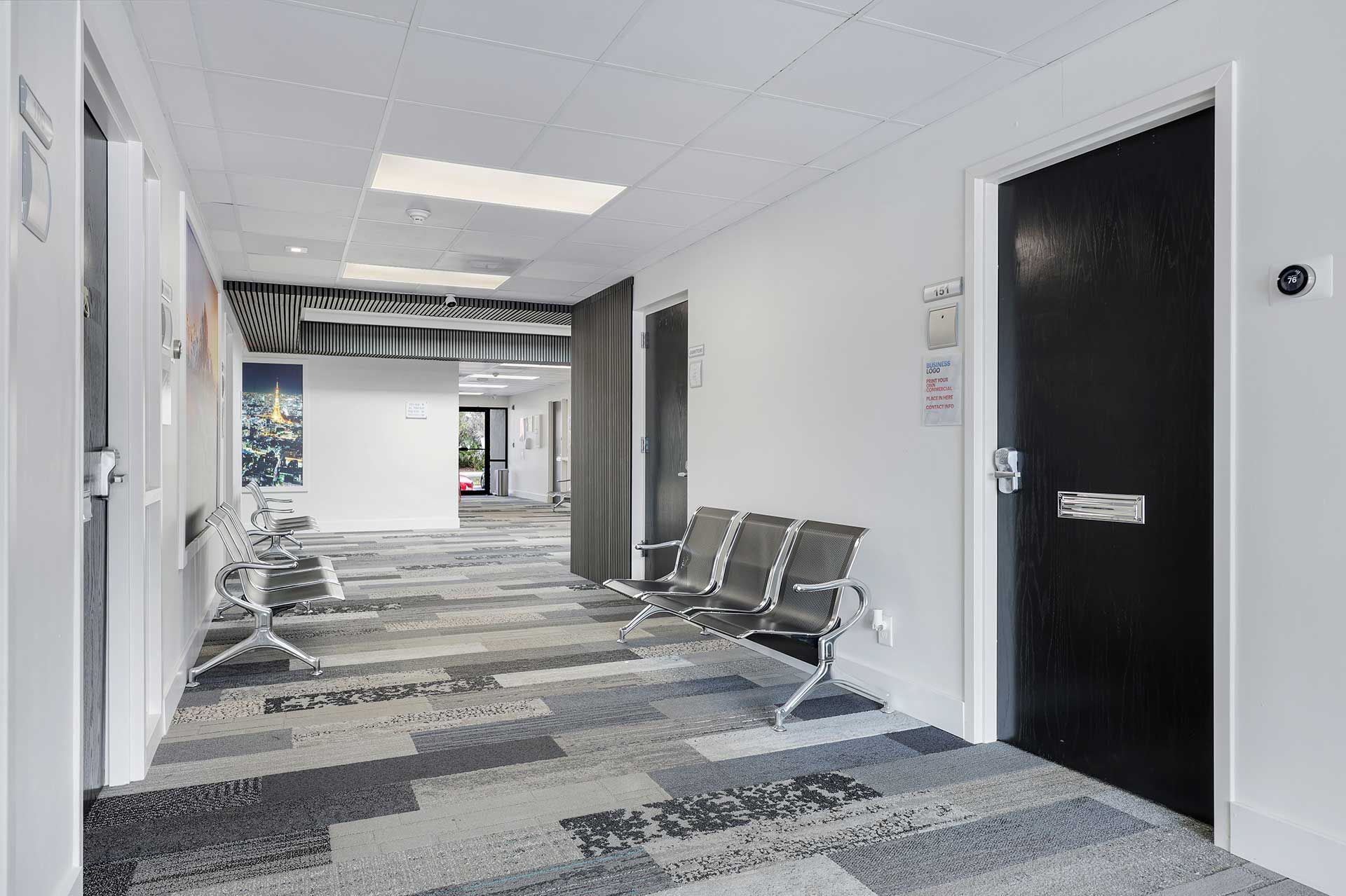 Hallway with waiting area: gray carpet, white walls, black doors, metal chairs.