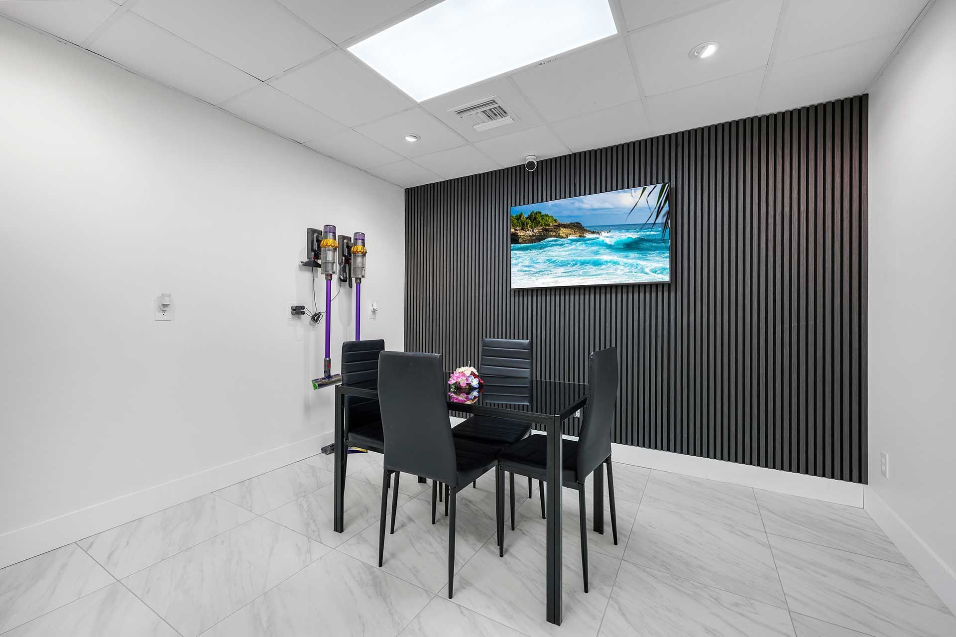 Small, modern office with a table, chairs, TV, and decorative wall. White walls, tile floor.