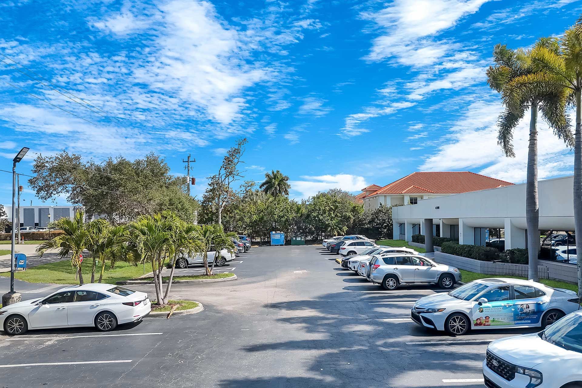 Parking lot with cars, trees, and buildings under a blue sky with clouds.