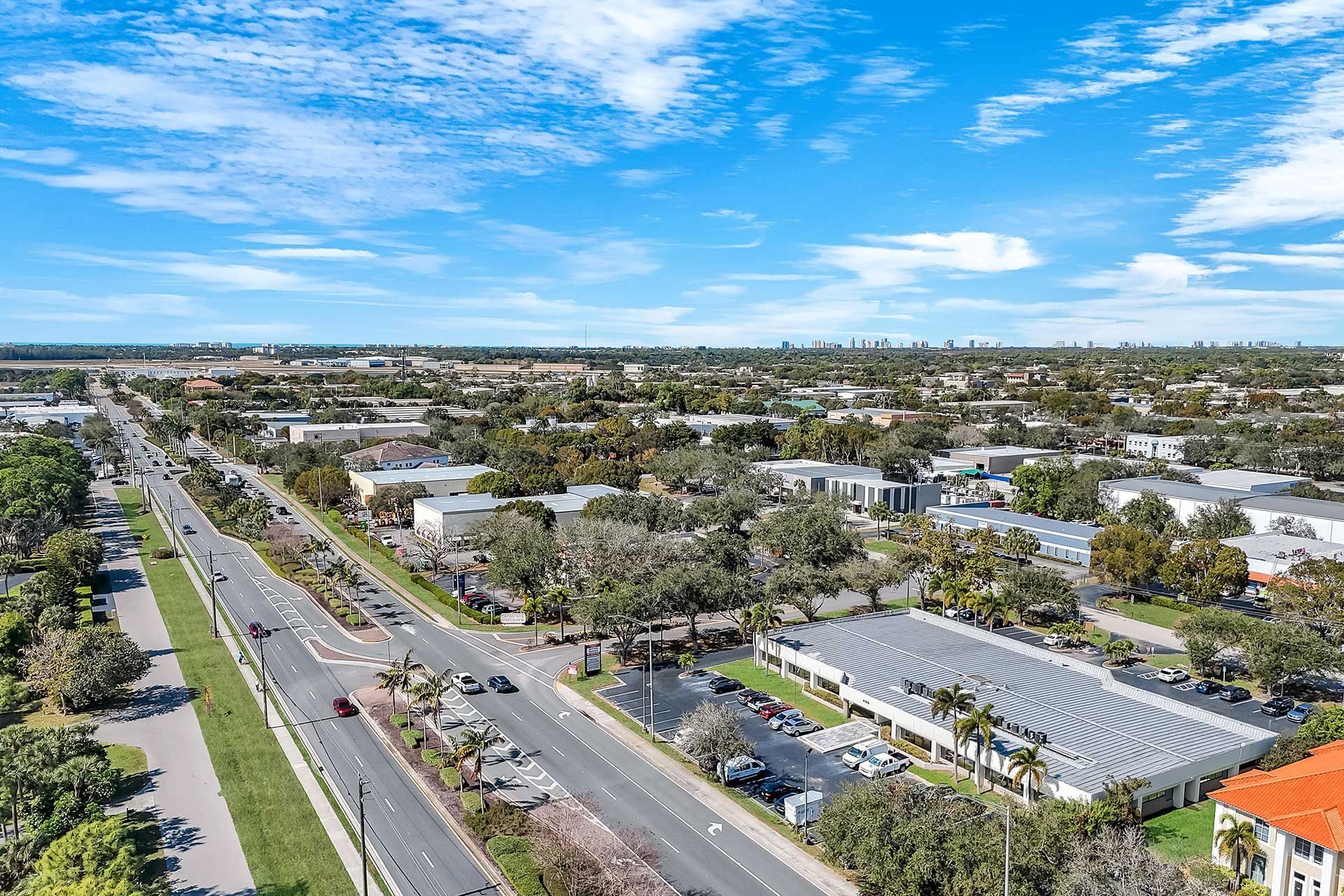 Aerial view of a city street lined with businesses, palm trees, and parked cars under a bright blue sky.