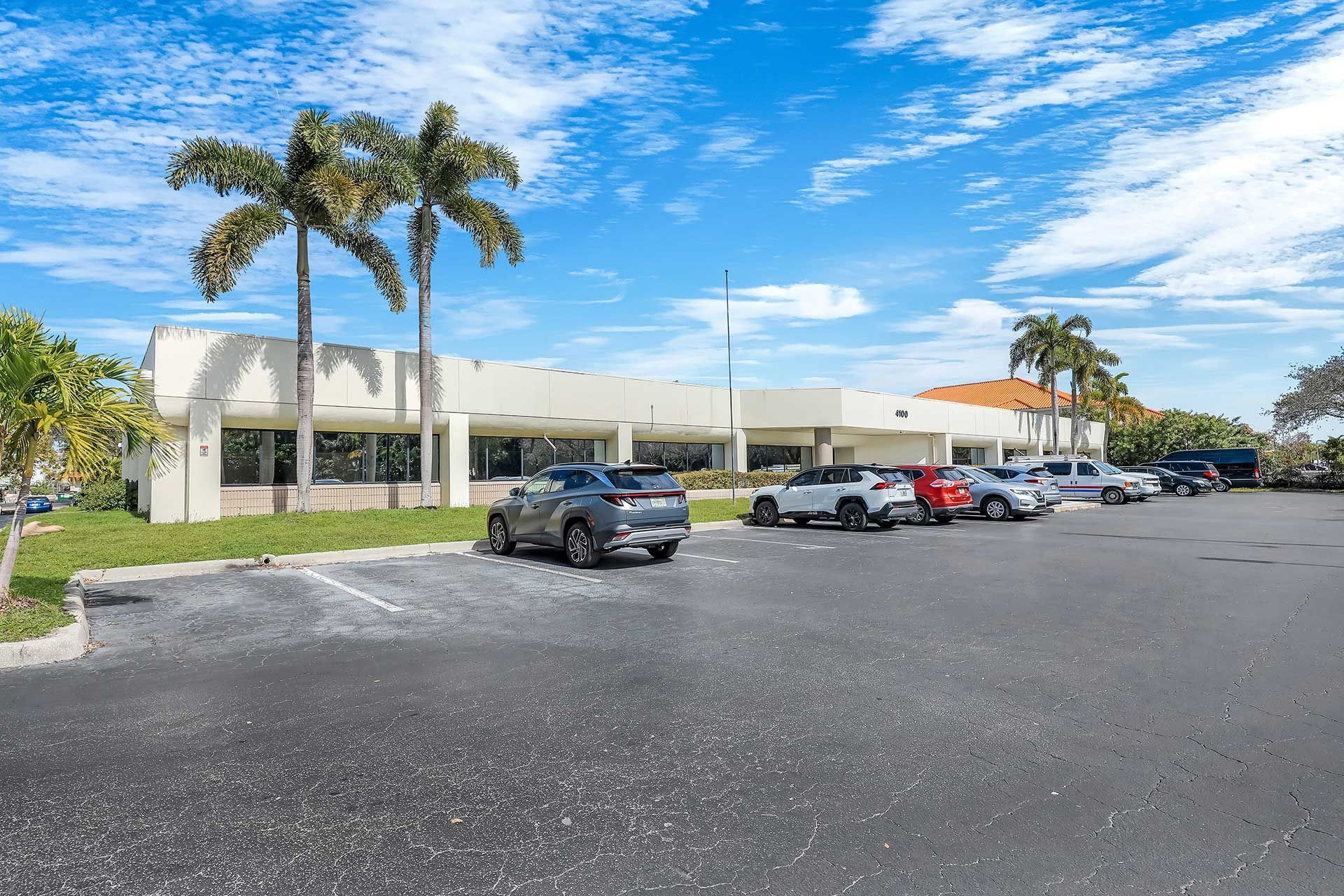 Parking lot in front of a low, white commercial building with palm trees and cars under a blue sky.