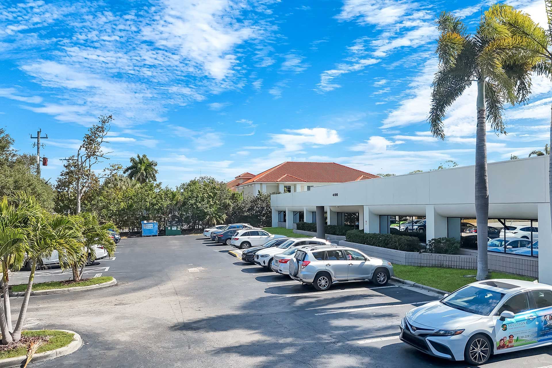 Parking lot with cars in front of a white building with an orange roof under a blue sky with clouds and palm trees.