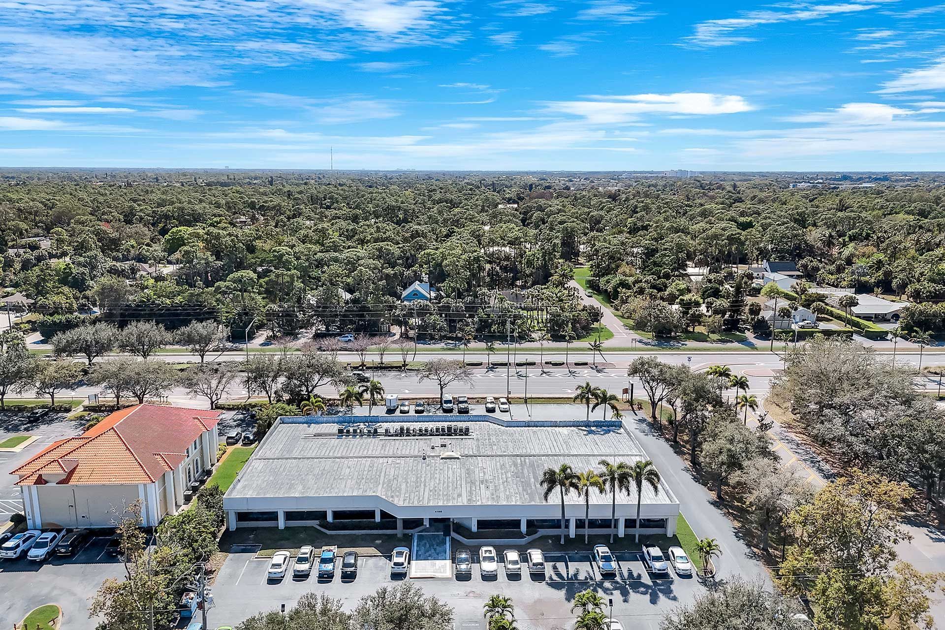 Aerial view of a white, low-rise building with a flat roof and parked cars in front, surrounded by trees under a blue sky.