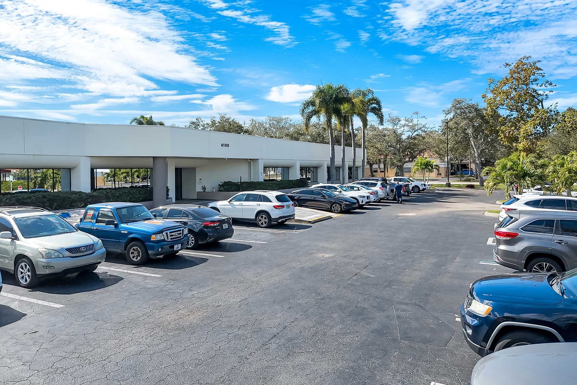 Cars parked in a black asphalt lot in front of a white building under a blue sky.