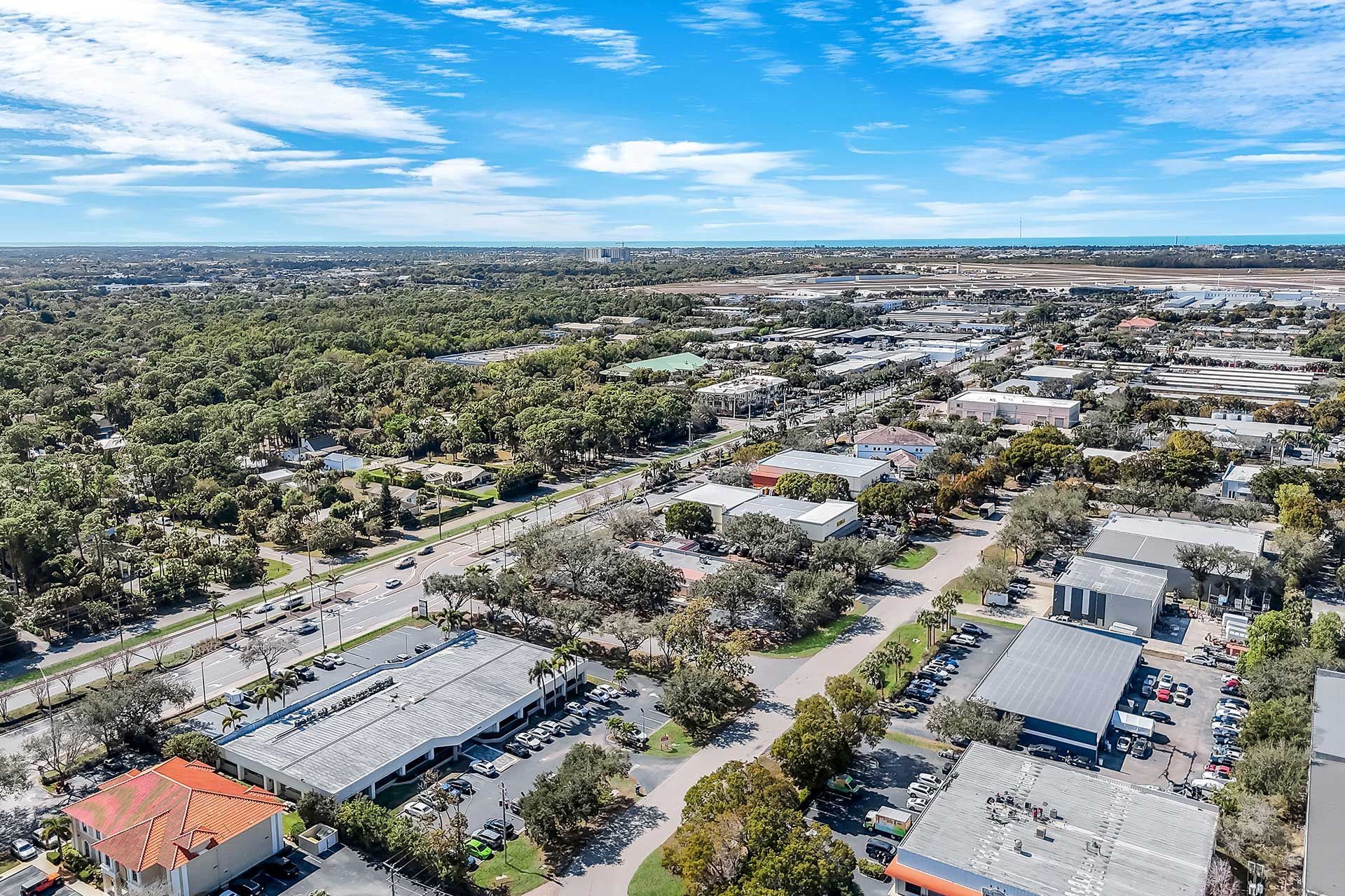 Aerial view of a town with trees, buildings, and a road under a blue sky with clouds.